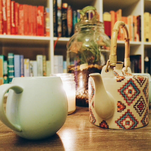 A mint mug of tea with a patterned teapot next to it, with a lit glowing candle and a terrarium in the background, against a backdrop of a colourful bookshelf