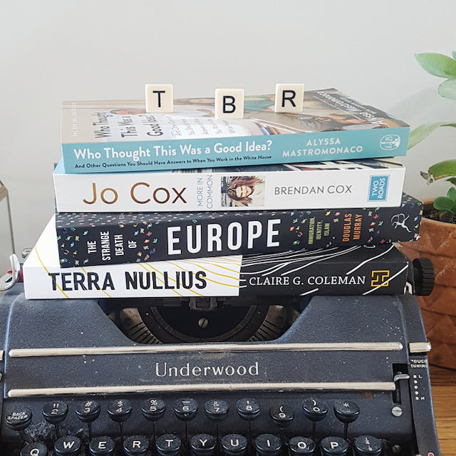 To Be Read TBR Pile of books stacked on top of each other, on top of an black Underwood vintage typewriter, with scrabble letters on top of the book stack with the singular letters 'T' 'B' and 'R'