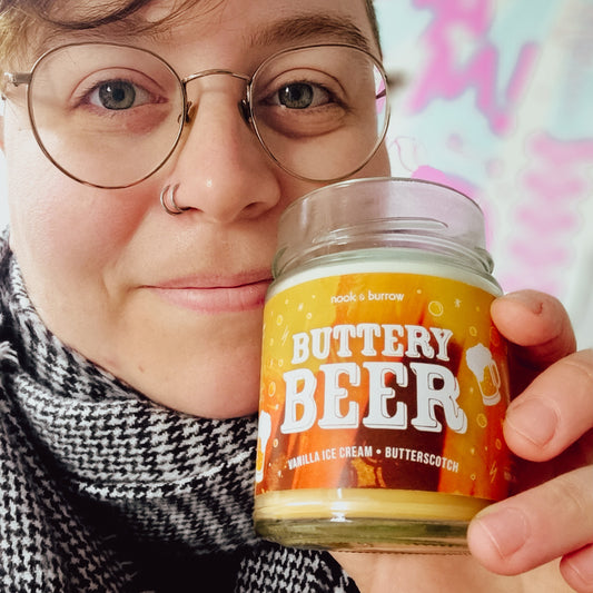 An androgynous person with a black and white scarf on and thin Harry Potter glasses holding a Buttery Beer candle up to their face
