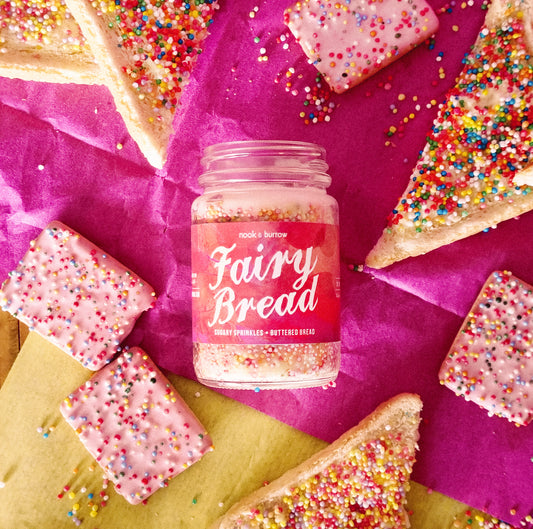 Fairy Bread candle with a pink label, surrounded by sprinkles and buttered bread with sprinkles on them and pink biscuits with sprinkles on them